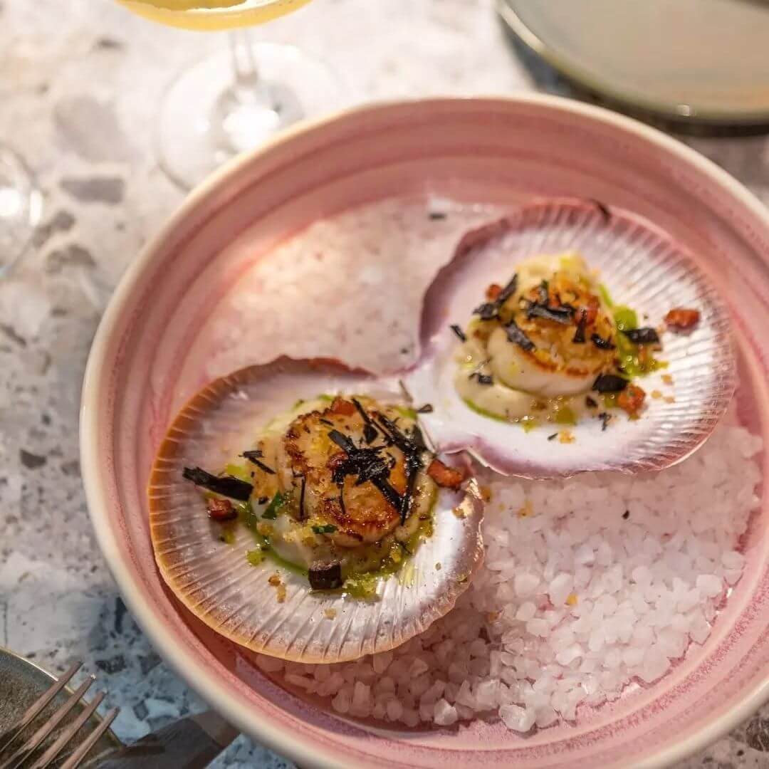 A dish with a bed of rocksalt topped with fresh scallops covered in a crumb